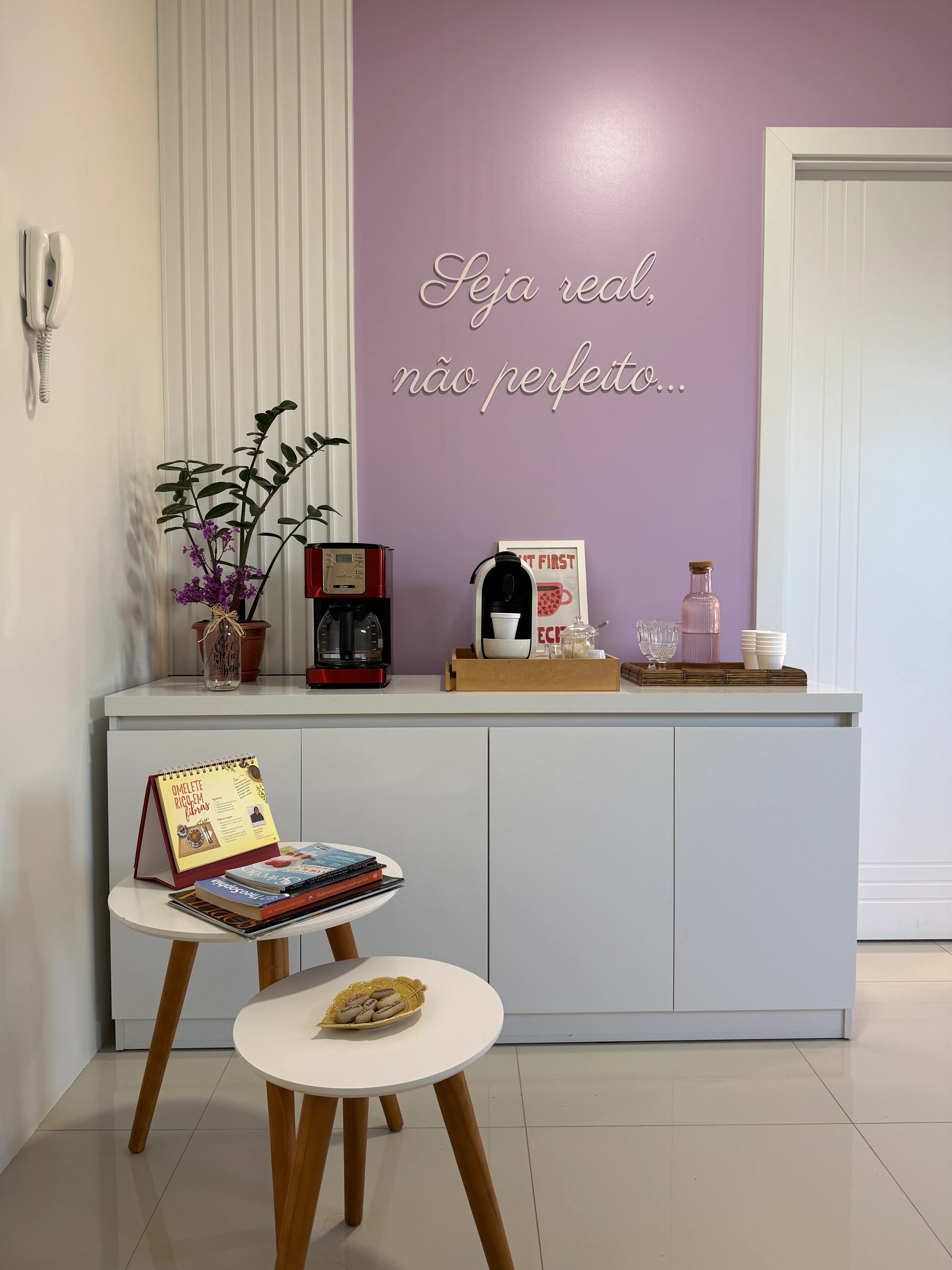 Buffet do café visto de frente, com cafeteiras, jarra rosa e a frase Seja real, não perfeito na parede